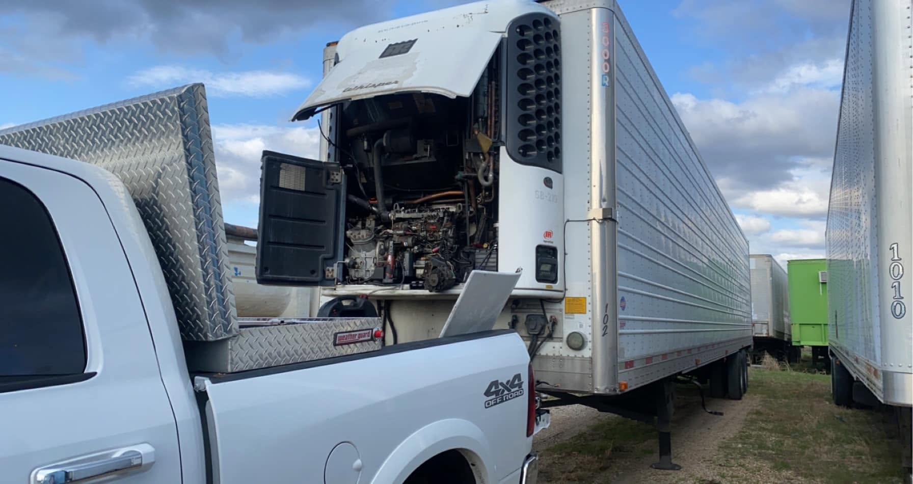 Reefer unit on a refrigerated trailer with its cover raised and engine exposed for on-site repair, service pickup truck parked alongside